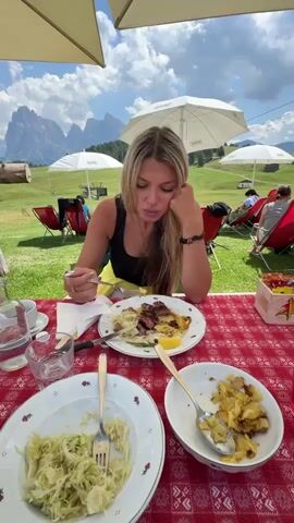 A woman with blonde hair is seated at a table with a red and white checkered tablecloth. She is wearing a black tank top and has her left hand resting on her cheek. In front of her is a plate of food, which she is eating with a fork. There are other plates of food on the table, including a dish of what appears to be shredded cabbage and a bowl of fried potatoes. In the background, there are several red chairs, umbrellas, and mountains. The sky is blue with some clouds.