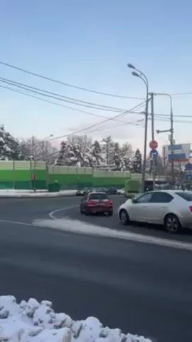 The video shows a winter scene at a road intersection. Cars are driving along the road, turning right and going straight. Snow is visible on the ground, especially on the sides of the road.  There is a green noise barrier and snow covered trees along the road.  A street lamp and utility lines are in the shot. Traffic signals are also visible.