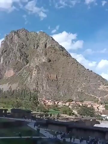The video shows a town nestled in a valley between large mountains. The town has buildings with red tile roofs and a structure with a grassy area and stone walls. There are people walking around the stone structure. The camera pans across the town and mountains, showing the valley and the surrounding landscape. The sky is blue with white clouds.