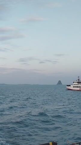 This media file captures a scenic view of the ocean with a ferry in the distance. The ferry, labeled "SEATRAN" in white letters on a red stripe, appears to be moving, indicated by subtle wakes in the water. The vessel itself is white with red accents and has multiple decks.

The setting is the ocean, with choppy, turquoise-blue water dominating the right side of the frame. The left side of the image is dominated by a pale, hazy sky, suggesting either early morning or late afternoon light. A faint, possibly mountainous landmass is visible on the horizon, behind the ferry. The overall atmosphere is calm and serene, with the soft light creating a peaceful mood. The presence of the ferry implies a transportation route or a journey. There are no people clearly visible in the media file. The perspective suggests the viewer is on another vessel or a platform at sea.