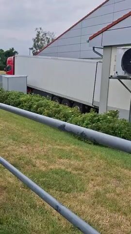 A white semi-trailer truck is parked next to a building with large industrial fans on the exterior. The truck is positioned at an angle, with its rear wheels close to a row of bushes. Beyond the truck and building, a street is visible with houses and trees. A blue car is driving on the street. The sky is overcast. The foreground shows a metal railing and grassy terrain.