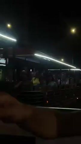 A night shot depicts a train filled with passengers riding past buildings. People are waving and smiling. Some passengers are looking at their phones. The train cars have red seats and black railings. The train is brightly lit with long white lights along the top. The buildings it passes are dark with lit windows.