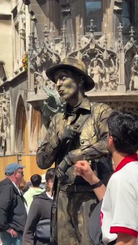 The video shows a street performer covered in gold paint, wearing a gold hat and clothing, standing still in a public square. The performer is interacting with a person in the foreground who is looking at them. Behind the performer is a large, ornate building with architectural details. In the background, there are other people walking around, some of whom are looking at the performer. The weather appears to be sunny, and the scene is set outdoors. The performer seems to be posing as a statue.