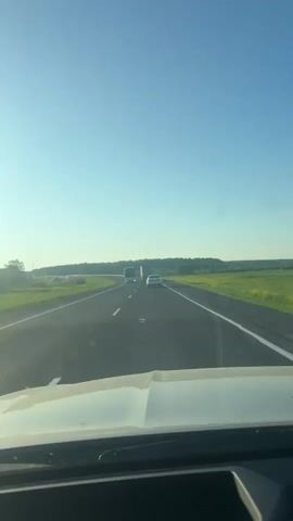 The video shows a first-person perspective from inside a vehicle traveling on a highway. The road is paved and has white lane markings. On the road ahead, there is a white car and a bus. The landscape on either side of the road is green and appears to be fields of grass and possibly some yellow flowers. The sky is clear and blue. The sun is shining, casting a bright light. The overall scene is one of driving on a rural highway on a sunny day.