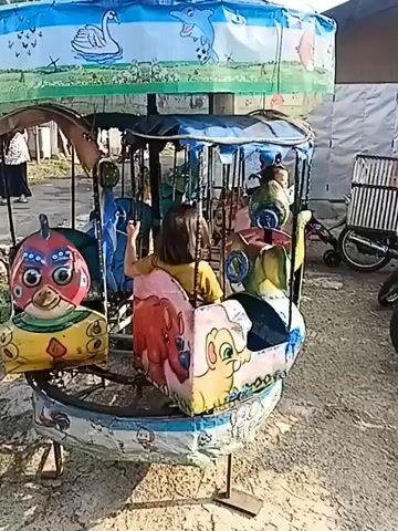 The video shows a children's carousel ride. A young girl with dark hair is seated in one of the cars, which is shaped like a pink rhinoceros. She appears to be looking to her right. Another child is visible in a car further back. The carousel has a colorful canopy with images of animals like an elephant, a swan, and dolphins. The ride itself has various animal-shaped seats, including a red bird and a blue dolphin. The carousel is outdoors, with a dirt or gravel ground visible. In the background, there is a motorcycle and a wheelchair. The carousel appears to be in use, as it is in motion.