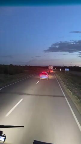 This media file depicts a view from inside a vehicle driving on a highway in Baiqadam, Kazakhstan, during twilight. The road is asphalt with clear white lane markings. In front of the vehicle, a silver car is driving in the same lane, its taillights illuminated. Further ahead, a large truck with a white trailer is visible. The sky is a deep blue, transitioning to a lighter hue near the horizon, with scattered clouds. The sun has set, casting a soft, ambient light. The surrounding landscape appears to be dry and somewhat barren, with low-lying vegetation and distant trees.

The activity is long-distance travel by car. The time of day is evening, indicated by the twilight sky and illuminated vehicle lights. The weather appears clear, with the exception of some clouds. There are two visible signs on the right side of the road, one with a blue background and white text, and another with a red circle and white symbols, likely traffic or directional signs. The overall mood is calm and transitional, as day turns into night. There are no discernible people in the frame, but the presence of vehicles indicates human activity. The emotional tone is peaceful, evoking a sense of journey and movement.