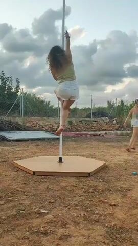 A woman with curly brown hair is performing a pole dance outdoors. She is wearing a light-colored tank top and white shorts. The pole is white and stands on a hexagonal wooden platform. The ground around the platform is dirt. In the background, there is a chain-link fence, some scattered rocks and debris, and green foliage. The sky is cloudy.