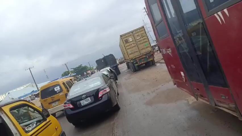 This media file depicts a traffic jam in Agbara, Nigeria. The scene is dominated by various vehicles, including a large red bus in the foreground, a container truck, a yellow minibus, and several cars and a tricycle. The road appears wet, suggesting recent rain, and the sky is overcast, indicating a cloudy day.

People are visible inside and around the vehicles. In the yellow minibus in the foreground, a person is seated in the driver's seat, and others appear to be passengers. In the background, a group of people are gathered near a building. The overall impression is one of congestion and slow-moving traffic, a common occurrence in urban or industrial areas.

Notable details include the distinctive yellow color of many of the vehicles, characteristic of public transport in Nigeria. The container truck suggests that the area is likely an industrial or logistics hub. There is some visible text on a sticker on the yellow minibus in the foreground, although it is too blurry to read clearly. The scene appears to be set during daylight hours. There are no overt indications of emotions, but the visual of a traffic jam inherently implies a degree of frustration or impatience for those involved. The setting, with its mix of vehicles, buildings, and power lines, points to a busy, functional, and possibly industrial area of Agbara, Nigeria.