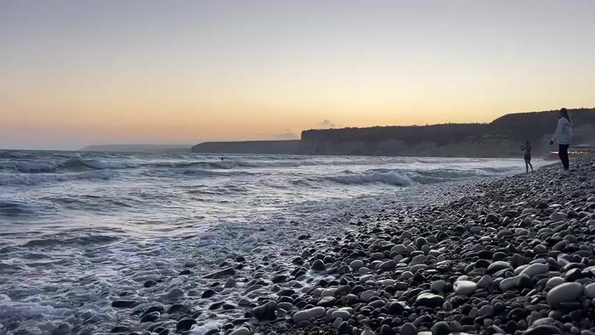 A low-angle wide shot shows a rocky beach with the ocean in the background. The waves are crashing on the shore. The sun is setting, casting a warm glow over the scene. A person is standing on the beach, looking out at the ocean. Another person is in the water, swimming. The sky is clear and the water is calm. There is a cliff face in the distance.