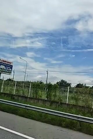 The video shows a view from a vehicle moving along a road. On the right side of the road, there is a metal fence topped with barbed wire, and behind the fence, there are trees and other vegetation. Further back, there are utility poles with wires. The sky is blue with scattered white clouds. The road itself has a white line marking the lane.