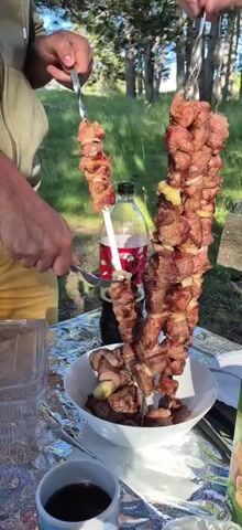 A person in a yellow shirt is using a fork and knife to remove cooked meat from a skewer. The skewer is made of metal and has chunks of meat and onions threaded onto it. The meat is browned and appears to be shish kebab. There are two large skewers of meat standing upright in a white bowl filled with more cooked meat. Behind the skewers is a clear plastic bottle of soda with a red label. The scene is outdoors, with green grass and trees in the background. The table is covered in aluminum foil, and there are other dishes and a glass on the table. The lighting suggests it is daytime, likely late afternoon due to the long shadows. The person's hands are visible and they are actively cutting the meat.