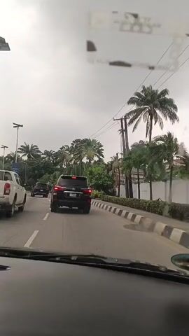 The video captures a view from inside a car, looking forward through the windshield. The car is driving on a paved road with distinct white lane markings. In front of the car, a black SUV is traveling in the same direction. Further ahead, another vehicle, possibly a white SUV, is also on the road. The environment appears to be tropical, with numerous palm trees and dense green foliage lining the roadside. Utility poles with wires are visible along the right side of the road. The sky is overcast, suggesting a cloudy day. The road has a curb with alternating black and white stripes. There is a slight reflection on the windshield that partially obscures the view.