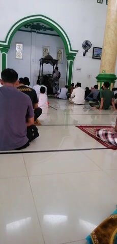 The video shows a group of men praying inside a mosque. They are all dressed in traditional Islamic attire and are facing the qibla, the direction of Mecca. The mosque appears to be simple and functional, with white walls, a tiled floor, and a few decorative elements. The men are performing the salah, the Islamic prayer, which involves a series of movements and recitations. The atmosphere is one of reverence and devotion.