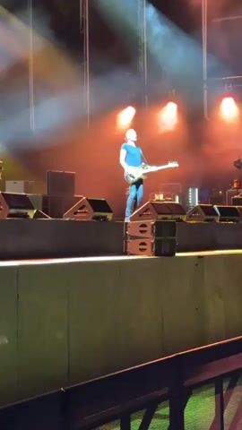 A man is playing a bass guitar on a stage. He is wearing a blue t-shirt, blue jeans, and dark shoes. He is standing on a stack of speakers. There are stage lights and smoke in the background. There are also other speakers and equipment on the stage. The stage is elevated above an audience area, and a railing is visible in the foreground.