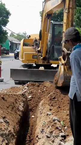 A yellow Komatsu excavator is digging a trench on a paved road. The excavator's arm is lowered into the trench, and the bucket is scooping dirt. A man wearing a light-colored shirt and a cap is standing next to the trench, looking at the excavator. Another person is visible inside the excavator's cab. To the left of the excavator, a white car is parked on the road. There are houses and trees in the background. The area appears to be an urban or suburban street undergoing construction.