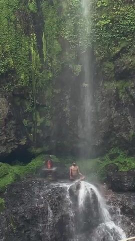 The video shows a man sitting in a meditative pose on a rock under a waterfall. The waterfall cascades down a rocky cliff covered in lush green vegetation. A small shrine-like structure sits to the left of the man, also on the rock. The water flows around and over the man and the rock. The environment is natural and verdant.