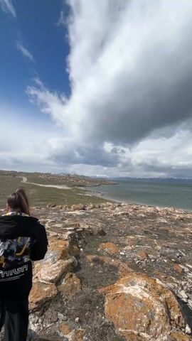 A person stands on a rocky landscape facing away from the camera, looking out towards a large body of water. The person is wearing a black hoodie and black pants. The hoodie has a large design on the back. The landscape is composed of rocks covered in orange lichen and patches of green grass. The water extends to the horizon, with a distant shoreline visible. The sky is partially cloudy, with patches of blue visible. A dirt road runs along the shoreline in the distance. The weather appears windy, as indicated by the movement of the clouds.