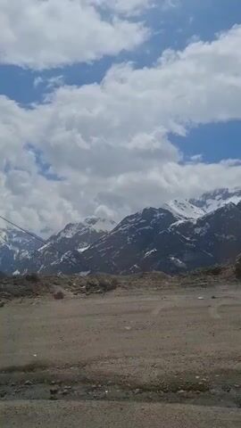 The video shows a mountain landscape on a cloudy day. In the distance, there are snow-capped mountains. A dirt road is in the foreground. A paved road runs along the side of a hill in the middle ground, and there are a few vehicles on it. There are power lines and a pylon. Some music plays in the background.