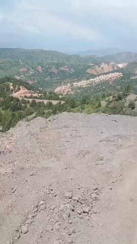The video shows a view from a high vantage point looking down into a valley. In the foreground, there is a rough, gravelly surface that appears to be a dirt path or a quarry. The ground is covered in small stones and dirt. Beyond the foreground, the terrain slopes down into a valley. The valley is filled with green vegetation, including trees and shrubs. There are also some patches of exposed earth with reddish-brown soil, suggesting a natural landscape or possibly some form of excavation or mining activity. In the distance, rolling hills and mountains are visible, with a hazy sky overhead. The overall impression is a natural, somewhat rugged landscape, likely in a mountainous or hilly region. The video appears to be a static shot with minimal camera movement, possibly taken from a stationary position.