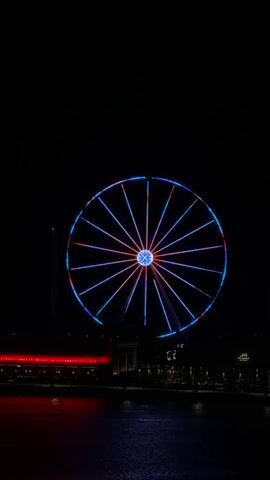This is a vertical, full-shot, nighttime video of a Ferris wheel. The Ferris wheel is illuminated with various colored lights that cycle through blue, pink, and red. The Ferris wheel is located in front of a dark background, likely a cityscape, with some buildings and lights visible below. In the foreground, there is a body of water reflecting the lights. The video captures the Ferris wheel rotating and the lights changing color, creating a dynamic visual display against the dark night sky. There are no people or interactions visible in the video.