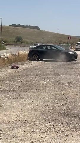 The video shows a red and black remote control car driving around a gravel lot. The RC car does a donut, kicking up dirt and dust. A dark sedan is parked with its front end in the grass on the left. A group of people walk into the scene in the background. In the middle of the scene, there is a dark SUV parked perpendicular to the camera. Trees and hills are in the background.