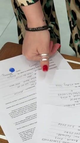 A person with red nail polish on their thumb uses a blue ink pad to thumbprint papers. The person is wearing three rings on the thumb and a bracelet on the wrist. The documents appear to contain emergency contact information. A leg is visible and appears to be wearing pants with a pattern of animal-like splotches. The surface beneath the papers is wooden.