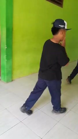A group of young people are walking down a hallway with green walls and tiled floors. The camera is positioned behind one of the individuals, a boy wearing a black t-shirt, blue pants, and a baseball cap. He walks with a slight limp, his left leg appearing to drag. Ahead of him, other individuals are visible, one in a yellow shirt and another in dark clothing. The hallway opens up to an outdoor area where more people are seen sitting. The boy in the foreground stops briefly, then continues walking, seemingly about to interact with the person in the yellow shirt.