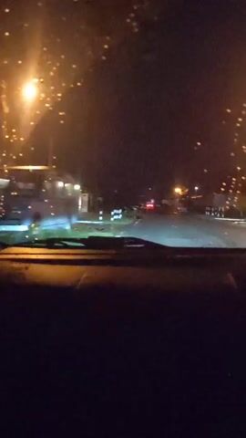 This is a first-person perspective video shot from inside a vehicle at night. The vehicle is moving forward on a road. The headlights of the vehicle illuminate the road ahead, which appears to be paved and has white lines marking the lanes. On either side of the road, there are reflective markers or bollards. In the distance, streetlights and the lights of buildings are visible. The sky is dark, indicating it is nighttime. There are also some blurred lights or reflections on the windshield. The scene appears to be in a semi-urban or suburban area, possibly with some businesses or residential buildings lining the road. No people are clearly visible.