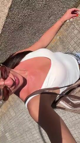 The video begins with a close-up of a woman lying down, her head slightly tilted. She is wearing a white tank top and denim shorts. Her skin appears tanned, and she has sunglasses on. The background is a textured wall and a tiled walkway. The camera then shifts to a wider shot, showing the woman walking, still wearing the same outfit. She is holding a bag and looking at the camera, smiling. The scene transitions to a view of a street with buildings and trees. The woman is now in focus, walking towards the camera and smiling. She has sunglasses on and a necklace. Her skin is visibly red, indicating a sunburn. The video ends with a shot of the woman walking away from the camera, with buildings and greenery in the background.