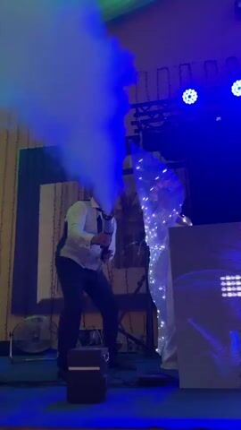 A man wearing a white shirt, black vest, black tie, and a hat operates a fog machine, directing smoke towards a person in a white outfit adorned with lights. The person in white stands behind a DJ setup with a large screen displaying colorful lights. The scene appears to be an indoor event with stage lighting, including blue and green lights. The camera pans and moves quickly throughout the clip, causing some blur.
