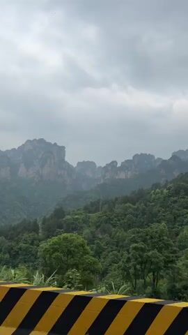 The video shows a view of mountains from a moving vehicle. The mountains are green with trees and have unique, tall rock formations on top. The sky is overcast, and the scene appears somewhat misty. In the foreground, there is a yellow and black striped barrier on the side of the road.