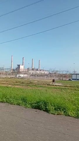The video shows a ride-along perspective from a vehicle moving along a road, passing by a large industrial facility. The sky is clear and blue. The facility includes several tall, cylindrical structures that appear to be smokestacks or chimneys, as well as an intricate network of electrical transmission towers and supporting structures. The ground is a mix of grass, bare earth, and gravel. The vehicle has a side mirror visible and is driving on a paved road with a guardrail. Power lines stretch overhead. The scene suggests a power plant or other large-scale energy production or distribution site.