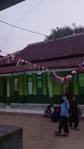 In an outdoor setting, a flag pole with a red and white Indonesian flag stands prominently in front of a building with a brown tiled roof and green and dark green walls. A string of red, white, and blue festive decorations hangs along the edge of the roof. Several children are present in the scene. In the foreground, a boy in a light blue shirt and dark pants stands near the flag pole, holding something upward. Another boy in a dark outfit is walking to the right. Several other children are seated or crouching on the ground to the left and in the background. The ground appears to be made of dirt or gravel. The sky is overcast, suggesting an early morning or late afternoon setting.