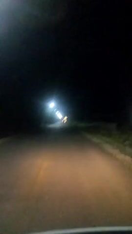The video shows a dimly lit road at night, likely captured from a moving vehicle. Headlights illuminate the asphalt ahead, creating a cone of light that dissipates into the darkness. Along the right side of the road, there appears to be a grassy embankment. In the distance, a bright light source, possibly a streetlight or a distant vehicle, casts a glow. Further down the road, a solitary cross is visible, illuminated by the same distant light. The overall atmosphere is one of a quiet, solitary journey through the darkness, with the cross serving as a point of focus or orientation.