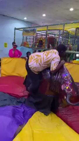 Here's a description of the video:

The video appears to be taken in an indoor amusement park or play center. The camera initially focuses on a man wearing a yellow patterned shirt and dark pants attempting to ride a mechanical bull, which is positioned on a cushioned surface of various colors. He struggles to maintain his balance.

In the background, other individuals are visible. To the left, a woman in a bright pink outfit observes. A large play structure with multiple levels and colorful elements is visible behind the man on the bull.

The camera then pans up and around, showing a wider view of the facility. The ceiling is decorated with modern light fixtures. Numerous people are present, including both children and adults. Some individuals are wearing uniforms, possibly staff. The area is filled with various arcade games and attractions, suggesting a bustling entertainment environment.