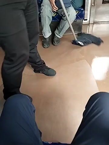 The video shows the interior of a train car. A person wearing black pants and black shoes is mopping the floor with a blue and white mop. Another person, wearing a black jacket and pants, is standing to the left, partially obscured. Two passengers are seated on a blue and white patterned bench in the background. The person in the foreground is wearing dark blue pants. The floor is a light brown, shiny surface that appears to be wet. The mop is moving across the floor, spreading the water.