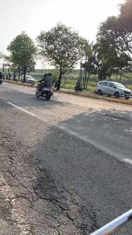 This is a short video clip filmed from a moving vehicle on a paved road, likely in Siring, Indonesia. The camera is positioned at a low angle, focusing on the road surface and the vehicles traveling in the same direction.

The primary subjects are several motorcycles and one white SUV. The motorcycles appear to be a mix of scooters and larger bikes, with riders dressed in casual clothing. The white SUV is a multi-purpose vehicle (MPV) and is prominently featured in the second half of the clip as it overtakes the camera's perspective.

The scene depicts a typical daytime traffic situation. The road is a two-lane asphalt surface with a clear white line marking the lanes. On either side of the road, there are trees with lush green foliage, suggesting a semi-rural or suburban environment. Beyond the trees, there are indications of fields or open land, and in the distance, some fencing and possibly infrastructure like poles or wires are visible.

The activity is simply traffic flow. Vehicles are moving along the road, and the camera itself is also in motion, creating a sense of forward progress. There are no obvious signs of any specific event or unusual activity.

Notable details include the time of day, which appears to be late afternoon or early evening, judging by the warm, soft sunlight casting long shadows. The weather seems clear and pleasant, with a bright sky. The location cues from the trees and the type of vehicles are consistent with a Southeast Asian setting. No strong emotions are conveyed in the footage, as it is primarily observational.

Visible text includes license plates on the vehicles. The SUV's license plate reads "AG 1225 LJ". Additionally, there is a logo on the rear of the SUV, though it is not clearly legible.