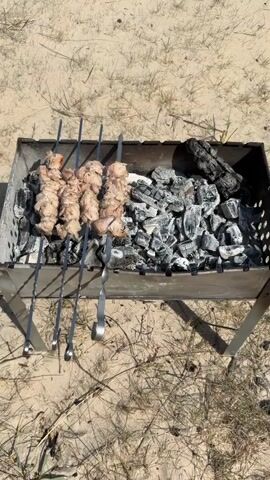 The video shows a sandy beach scene. In the foreground is a metal grill with skewers of meat cooking over charcoal. To the right of the grill, some unburnt wood logs are visible. Moving away from the foreground, a woman is sitting in a green camping chair, holding a glass. She is wearing a pink shirt and tan pants, and her hair is in a braid. Further in the background, a young girl in a pink outfit is blowing bubbles near the water's edge. The ocean is visible in the distance under a blue sky with sparse clouds.