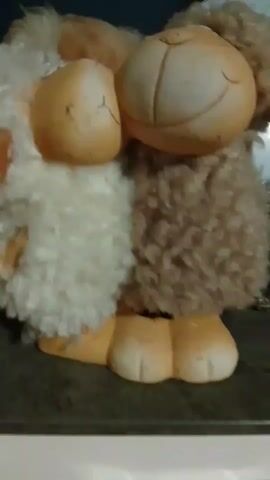 The video is a close-up shot of a stuffed animal, likely a sheep or lamb, on a dark surface. The stuffed animal is predominantly tan and white, with fluffy, curly tan fur around its neck and head. Its body appears to be made of a softer, white material, with tan hooves. The stuffed animal is positioned facing slightly to the left, with its head tilted. The lighting is dim, and the background is out of focus, showing a hint of a white object and some green and white patterns, possibly part of a clock or another decorative item. The camera is moving slightly, causing some blur, particularly in the fur and background.