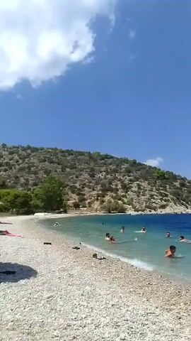 The video pans across a pebbled beach with clear turquoise water lapping at the shore. The sea is a deep blue further out, with a small island in the distance. Several people are swimming and enjoying the water. On the left side of the frame, a green hill covered in dry vegetation rises steeply from the beach. The sky is a vibrant blue with scattered white clouds. In the distance, a few boats are visible on the water, and buildings are scattered on the hillsides.