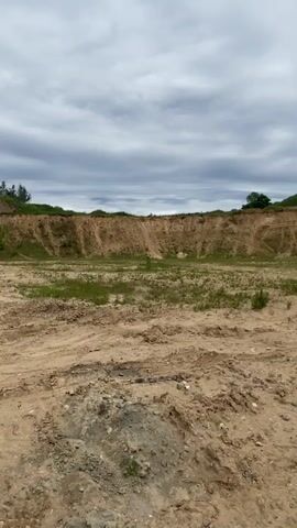 The scene is a wide shot of a sand pit on an overcast day. The pit has high sandy embankments on three sides and is mostly barren with patches of low green vegetation. The ground in the foreground is sandy and rough.  A single person on a dirt bike is visible towards the back of the pit, riding from right to left.