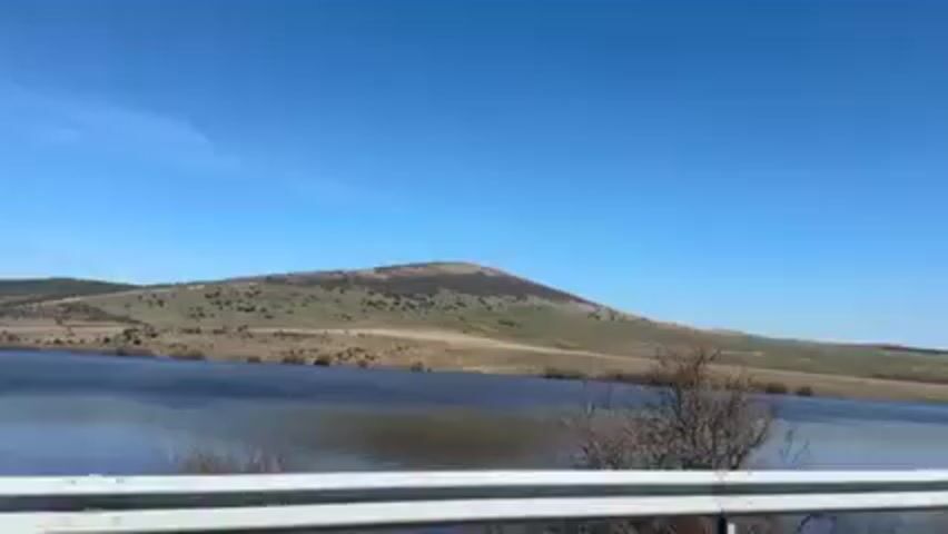 The video shows a view of a lake with a small hill in the background, under a clear blue sky. The lake's surface is slightly disturbed by the wind. On the hill, which is primarily covered in grassy vegetation, several wind turbines are visible. Some leafless bushes are near the lake. A metal guardrail is in the foreground.