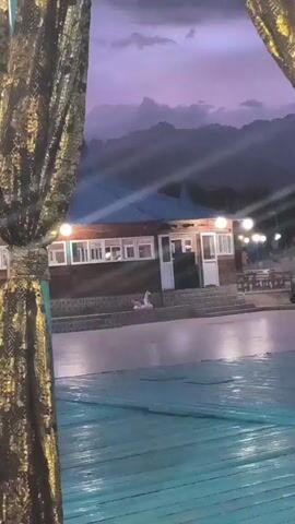 The video opens with a view of a wooden building with a blue roof, framed by ornate curtains. In front of the building, there's a large paved area, possibly a patio or an empty pool, with a pink flamingo inflatable toy on the ground. The sky above is a dusky purple with clouds, and mountains are visible in the distance. Lights are on at the building, illuminating the entrance and windows. As the video progresses, a red light briefly flashes on the building's exterior. The overall atmosphere suggests an evening setting, possibly at a resort or outdoor venue.
