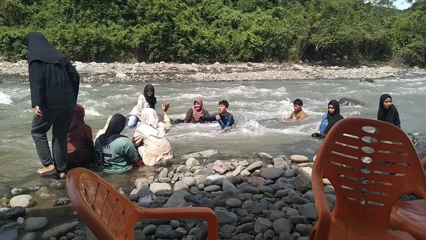 This media file depicts a group of people enjoying a day at a river in Meurah, Indonesia. The scene is set by a flowing river with a rocky shore. Lush green trees and vegetation line the background, suggesting a natural, possibly rural, setting. The weather appears to be sunny, casting bright light on the water and surrounding landscape.

Several individuals, including women in hijabs and young men, are in or near the water. Some are sitting on the river rocks, while others are wading or playing in the moderately flowing water. One young man is actively splashing in the river, with others around him reacting to his playful actions. The overall atmosphere seems relaxed and joyful, with the people enjoying a recreational activity outdoors.

In the foreground, two orange plastic chairs are visible, implying a place where people might sit to rest or observe. The rocky riverbed is prominent, with many smooth, grey stones of various sizes. The water itself is somewhat murky, typical of a natural river. The time of day is likely midday, given the brightness of the sunlight and the shadows. There is no visible text in the media file.