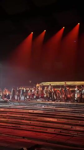 The video captures a live theatrical performance on a large, multi-level stage, likely indoors at night. The stage is dimly lit, with dramatic red and white spotlights illuminating the performers and set pieces.

The initial shot shows a large ensemble of performers, dressed in period-appropriate costumes, standing in rows on wide stone-like steps that form part of the stage. Red spotlights cast long, downward beams over them.

A brief pan then reveals more of the stage and a bright white spotlight.

The final, extended shot focuses on a prominent wooden boat with a large, red, rectangular sail, resembling a traditional Chinese junk, positioned center stage. Several performers are on board this main boat, while others are arranged around it in smaller wooden boats or on the stage platforms. One performer near the front holds a torch, emitting a warm glow. The stage floor and steps leading up to the boat are also illuminated with reddish light, suggesting firelight or a dramatic evening scene. The overall atmosphere is grand and theatrical, depicting a historical or epic narrative, possibly set in China, given the location context of Shapingba District.