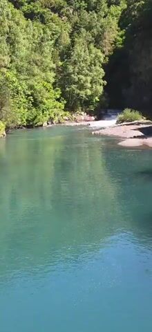 The video shows a clear blue-green river flowing towards a small waterfall. Lush green trees and foliage line the banks of the river, with some rocks and gravel visible along the edges. The sun is shining, casting light and reflections on the water. The waterfall is cascading down a rocky incline, creating white foam as it flows into the river below. The water appears calm and serene in the foreground, contrasting with the gentle movement of the waterfall.