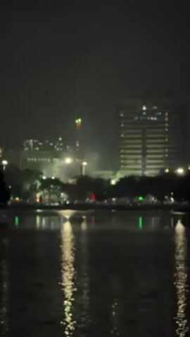 At night, the video reveals a cityscape reflected in a body of water, with fireworks intermittently lighting up the dark sky. The water's surface mirrors the lights of the city, which includes several buildings, one of which is a tall, well-lit tower. Trees line the water's edge, interspersed with streetlights. The fireworks burst irregularly, momentarily brightening the scene before fading into the night.