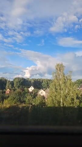 The video shows a view of a rural landscape. There are trees and houses visible, as well as a cloudy sky with a faint rainbow. The camera is panning across the scene, providing a sweeping view of the area. The lighting suggests it is daytime.