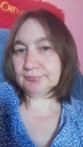 A woman with shoulder-length brown hair and a blue patterned shirt is shown from the chest up. She has a small smile and appears to be looking towards the camera. Behind her, a pink background is visible with a red plush object with yellow lettering that says "Oingo Boingo". A blue patterned pillow is also visible behind her.