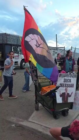 The video shows a group of people gathered outside, some standing and some sitting, near a chain-link fence topped with barbed wire. A person with pink hair and red and blue leggings is sitting on the ground in the foreground.  Next to them is a green wagon carrying various items, including an orange crate and a sign that reads "Mushroom Dick Messiah" above a picture of Donald Trump. Attached to the wagon is a large pride flag with a black fist on it.  Other people are standing around, including one wearing headphones and a man with a camera. In the background, there is a white truck with "National" printed on it. The sky is visible above, with a mix of blue and sunset colors. Some people are holding signs.